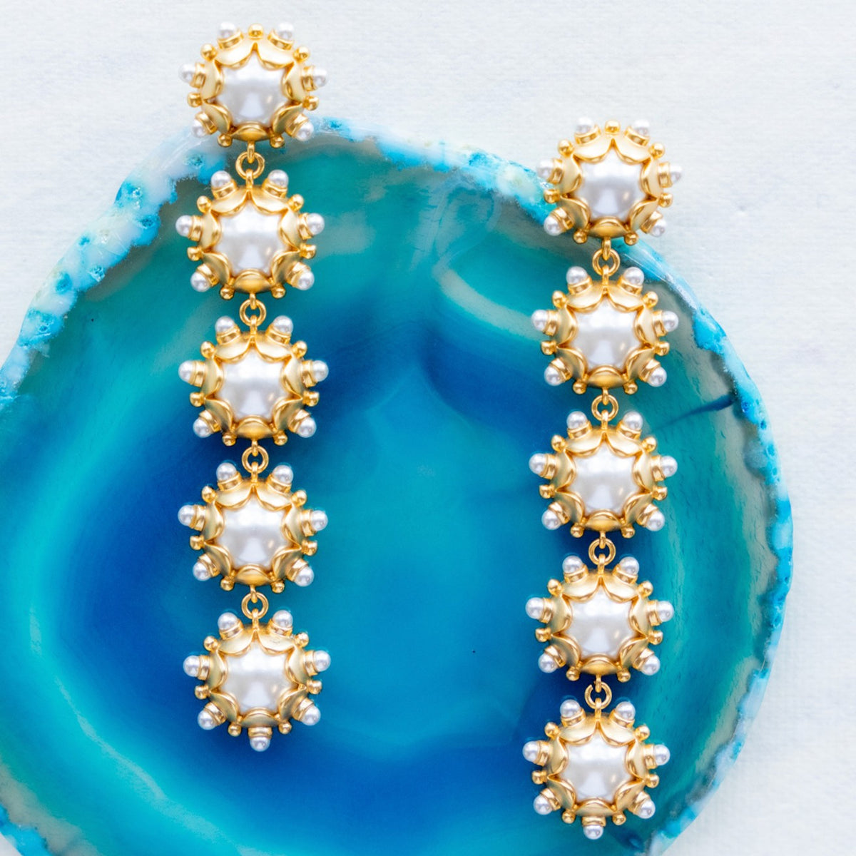 Gold and pearl Perla drop earrings arranged on a blue agate slice