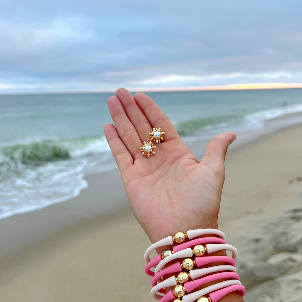 Hand wearing gold rings and a pink silicone bracelet stack beside the ocean